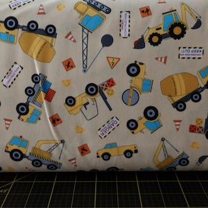 May include: Beige fabric with a repeating pattern of yellow construction vehicles, including a crane, cement mixer, bulldozer, and a road roller. The fabric also features yellow and black construction signs, including "Danger", "Keep Out", and "Under Construction".