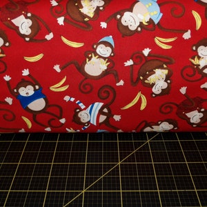 May include: Red fabric with a repeating pattern of monkeys in various poses. The monkeys are brown with blue and white accents. Some monkeys are wearing hats. The background is red with yellow banana shapes.