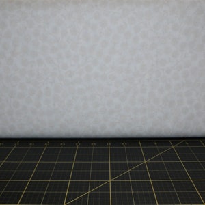 Puede incluir: Una tela blanca con un sutil estampado floral en toda la superficie. La tela se muestra sobre una alfombra de corte negra.