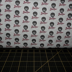 May include: A white fabric with a repeating pattern of Bob Ross's face and the words "Bob Ross" in red. The fabric is folded in half and the bottom edge is visible.