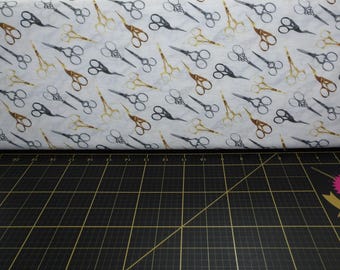 May include: White fabric with a repeating pattern of gold and silver scissors. The fabric is folded in half and lying on a black cutting mat.