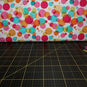 Puede incluir: Tela blanca con un patrón de lunares de colores. Los lunares son de varios tonos de rosa, naranja, turquesa y verde. La tela está doblada por la mitad y sobre una estera de corte negra.