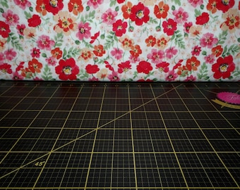 Puede incluir: Una tela blanca con un patrón floral con flores rojas, rosas y naranjas. La tela está doblada por la mitad y colocada sobre una estera de corte negra.