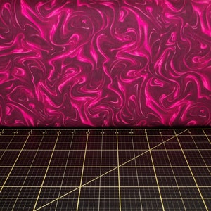 May include: A close-up of a pink and purple fabric with a swirling marble pattern. The fabric is lying on a black cutting mat with a ruler and a sewing needle.