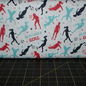 May include: White fabric with a repeating pattern of blue, red, and teal silhouettes of soccer players. The fabric has the text "Play Like A Girl" repeated throughout.