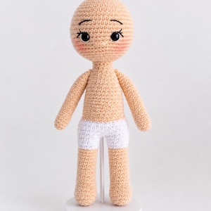May include: A crochet doll with light peach yarn skin, black embroidered eyes, and pink blush on the cheeks. The doll wears white crocheted shorts and is supported by a white vertical stand with a circular base, standing upright on a plain surface.