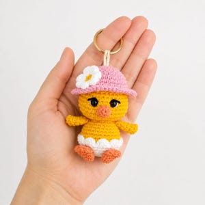 May include: A handmade yellow crochet chick keyring wearing a pink hat with a white flower. The chick has orange feet and a white trim. The keyring has a gold-coloured ring.