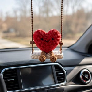 May include: A red crocheted heart-shaped ornament with a smiling face, sitting on a small swing. The swing is suspended by brown cords, designed to hang from a car's rearview mirror. The background is blurred, suggesting an outdoor setting.