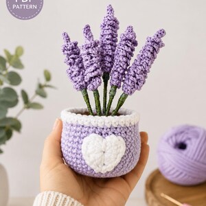 Puede incluir: Una planta de lavanda de ganchillo en una maceta de color violeta claro con una aplicación de corazón blanco. La planta presenta cinco tallos de flores moradas texturizadas sobre tallos verdes. Un sello circular contiene el texto PDF PATTERN.