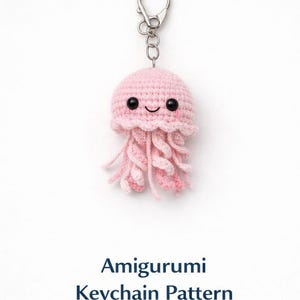 Könnte beinhalten: Ein rosa gehäkelter Quallen-Schlüsselanhänger mit einem silbernen Verschluss. Die Amigurumi-Qualle hat ein lächelndes Gesicht und fließende Tentakel. Der Text "Amigurumi Keychain Pattern" steht darunter.
