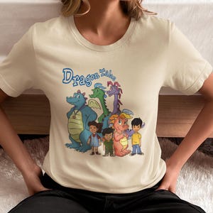 Op de afbeelding: Crèmekleurig T-shirt met de tekst "Dragon Tales" in blauw schrift boven een cartoonillustratie van draken en kinderen. De draken hebben verschillende kleuren, waaronder blauw, groen en paars. De kinderen dragen verschillende outfits.