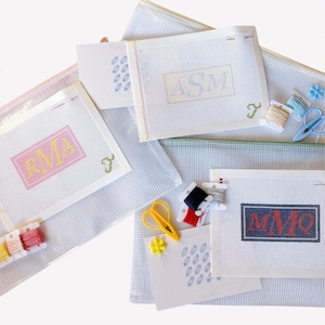 May include: Needlepoint canvases with personalized initials in pink, yellow, and navy blue. The canvases are accompanied by embroidery floss, scissors, and other needlepoint supplies. The background is a light blue and white grid pattern.