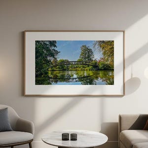 May include: A framed landscape print featuring a wooden arched bridge over a calm lake, surrounded by lush green trees and foliage. The water reflects the bridge and the sky. The artwork is displayed in a living room setting.