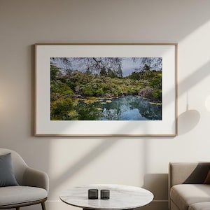 May include: A framed landscape photograph depicts a serene pond surrounded by lush greenery and trees. The artwork is displayed on a neutral-colored wall, complemented by modern furniture and decorative lighting fixtures, creating a calming atmosphere.