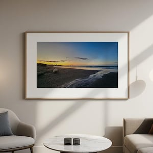 May include: A rectangular landscape photograph in a light wood frame featuring a sunset over a sandy beach with a small stream flowing toward the ocean. The sky transitions from deep blue to orange and yellow hues above the dark shoreline.