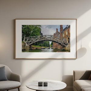 May include: A framed print of a wooden bridge over a river, with a brick building in the background. The bridge has an intricate design and is surrounded by lush greenery. The artwork is displayed in a living room setting.