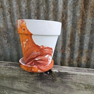 May include: White ceramic flower pot with a decorative orange and brown abstract design. The pot sits on a matching saucer. The pot is displayed on a wooden surface with a corrugated metal background.
