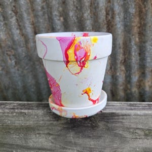 Op de afbeelding: Witte keramische bloempot met bijpassende schotel, versierd met een levendig abstract ontwerp in roze, geel en rood. De pot heeft een klassieke vorm, perfect voor het planten van bloemen of kruiden. De pot is ongeveer 10 cm hoog.