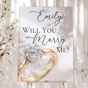 Könnte beinhalten: Ein Schild auf einer silbernen Staffelei mit dem Text "Emily Will You Marry Me?" und einer Aquarellillustration eines goldenen Verlobungsrings mit einem großen Diamanten. Das Schild befindet sich vor einem weichen, weißen Hintergrund.