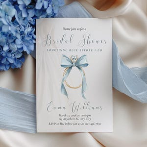 Op de afbeelding: Een uitnodiging voor een bruidsdouche met een aquarelontwerp met een ring met een blauwe strik en diamant. De uitnodiging bevat de tekst "Bridal Shower" en evenementdetails. Blauwe hortensia's en een lint accentueren de uitnodiging.