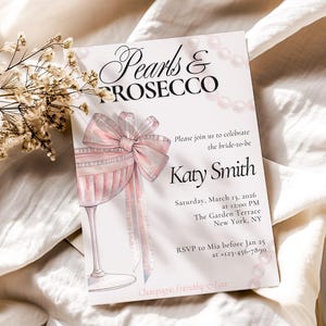Op de afbeelding: Een witte uitnodigingskaart met de tekst "Pearls & Prosecco" en "Katy Smith". De kaart toont een aquarel illustratie van een champagneglas met een roze strik en parels. De uitnodiging is voor een bruidsevenement.