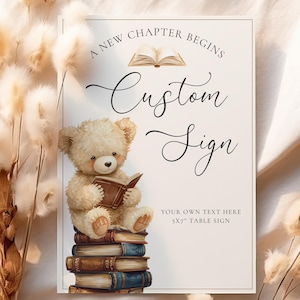 Op de afbeelding: Een rechthoekig aangepast bord met de tekst "A New Chapter Begins" en "Custom Sign". Een teddybeer zit op een stapel boeken en leest een boek. Het bord is een tafelbord van 12,7 x 17,8 cm.