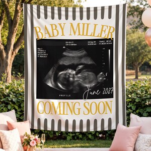 Può includere: Uno striscione rettangolare in tessuto con bordo a righe verticali bianche e nere, testo dorato BABY MILLER e COMING SOON e un'ecografia centrale con il testo scritto a mano June 2027 stampato sulla superficie.