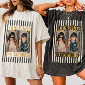 Op de afbeelding: Twee oversized t-shirts, één crème en één antracietgrijs, elk met een centrale afbeelding met verticale strepen. Het ontwerp bevat de tekst "LOOK WHO'S GOT MARRIED" in goud en een foto van twee kinderen die het vredesteken maken.
