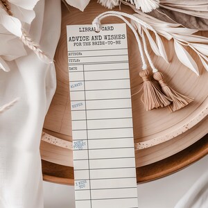 Könnte beinhalten: Eine weiße Bibliothekskarte mit dem Text "ADVICE AND WISHES FOR THE BRIDE-TO-BE" in Schwarz gedruckt. Die Karte hat Linien für Autor, Titel und Datum, mit Aufforderungen wie "ALWAYS" und "NEVER". Sie ist an einer weißen Schnur mit zwei beigen Quasten befestigt.