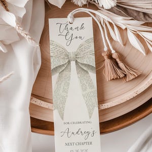 May include: A bookmark with a bow design made from book pages. The bookmark has the words "Thank You" at the top, and "For Celebrating Audrey's Next Chapter 05.26.2026" at the bottom. It has a white cord and two tassels.