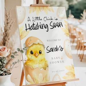 Puede incluir: Un letrero de baby shower sobre un caballete de madera. El letrero presenta el texto "A Little Chick is Hatching Soon" y "Welcome to Sarah's Baby Shower 05.05.2027". Una ilustración en acuarela de un pollito amarillo saliendo de un huevo está en el centro.