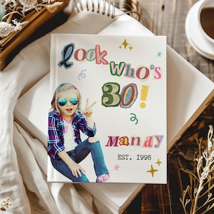 May include: A white hardback book featuring a collage-style cover with the text "look who's 30!", "MANDY", and "EST. 1996". The cover includes a photograph of a child wearing sunglasses and a plaid shirt, accented by gold star graphics.