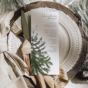 Pode incluir: Um cartão de menu de Natal com um design de árvore de Natal em aquarela, colocado em um cenário de pratos em camadas. A configuração inclui talheres dourados, um guardanapo de linho e elementos decorativos, criando um arranjo de mesa festivo elegante.