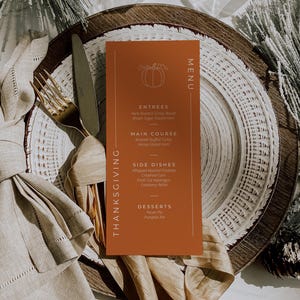 May include: Thanksgiving menu card on a plate with a fork and knife. The menu is orange with white text and a pumpkin illustration. The menu lists entrees, main course, side dishes, and desserts. The plate is on a table with a napkin and decorative elements.