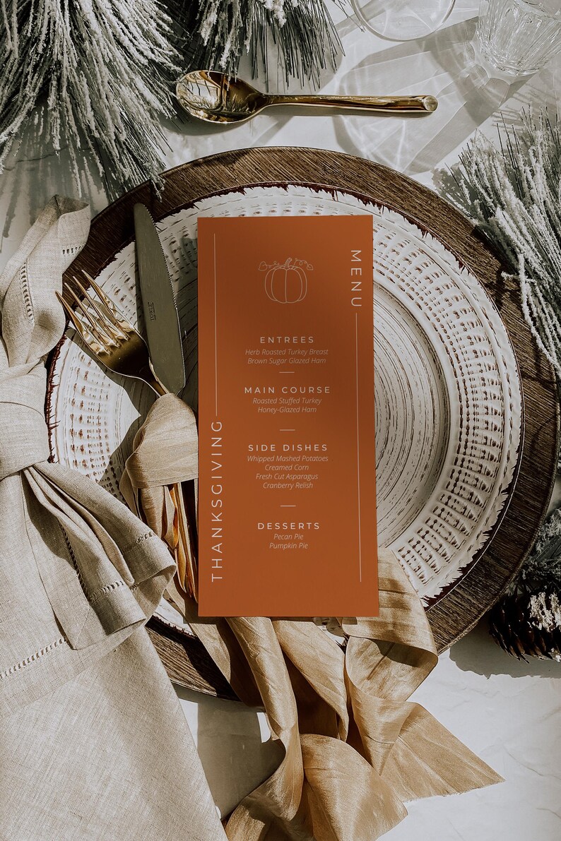 May include: Thanksgiving menu card on a plate with a fork and knife. The menu is orange with white text and a pumpkin illustration. The menu lists entrees, main course, side dishes, and desserts. The plate is on a table with a napkin and decorative elements.