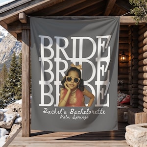 A grey custom bachelorette banner hangs on a wooden porch against a mountain backdrop, displaying stacked white BRIDE text and a child photo with Rachel's Bachelorette Palm Springs text.