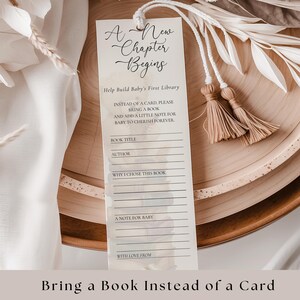 Op de afbeelding: Een bladwijzer met de tekst "A New Chapter Begins" en "Bring a Book Instead of a Card". De bladwijzer heeft ruimte om de titel van het boek, de auteur en een notitie voor de baby te schrijven. Het is vastgebonden met een wit koord en heeft twee beige kwasten.
