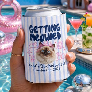Puede incluir: Un enfriador de latas de espuma con rayas azul claro y blancas que presenta un gráfico de un gato con una corona rosa. El texto dice Getting Meowied, Kate's Bachelorette y Charleston, 2026, junto a ilustraciones rosas de una bola de discoteca.