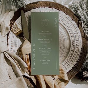 May include: A Thanksgiving menu card in sage green with a pumpkin illustration, placed on a layered plate setting. Gold cutlery and a linen napkin complement the autumnal theme. The menu lists entrees, main course, side dishes, and desserts.