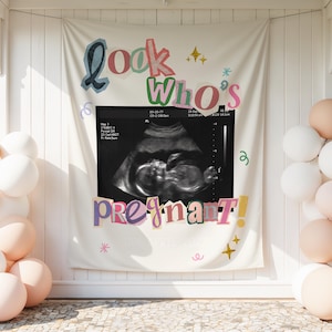Può includere: Uno striscione in tessuto color crema con la scritta "Look Who's Pregnant!" in lettere colorate e giocose. Al centro dello striscione è presente un'ecografia in bianco e nero di un feto. Lo striscione è affiancato da grappoli di palloncini bianchi e pesca.