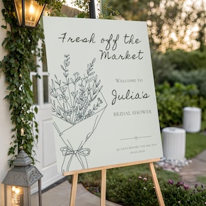 Op de afbeelding: Een rechthoekig lichtgroen bord op een houten ezel met een zwarte lijntekening van een bloemenboeket. De tekst luidt: Fresh off the Market, WELCOME TO Julia's BRIDAL SHOWER, 45 DAYS BEFORE THE BIG DAY en 01.01.2026 met een klein hartje.