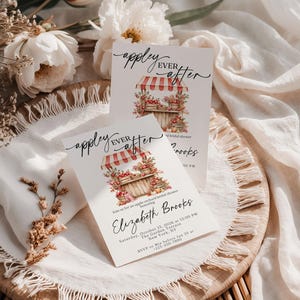 Puede incluir: Dos tarjetas de invitaci&oacute;n rectangulares blancas con el texto "appley EVER after" en caligraf&iacute;a negra. Las tarjetas muestran una ilustraci&oacute;n en acuarela de un puesto de manzanas de madera con un toldo a rayas rojas y blancas, lleno de manzanas.