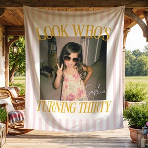 May include: A rectangular fabric banner featuring a light pink and white vertical striped background. The text LOOK WHO'S TURNING THIRTY is printed in gold. A central photograph shows a child wearing sunglasses, and the name Sophia is written in script.