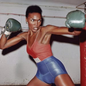 May include: A person wearing a red vest top and blue shorts, boxing with green gloves. The person is in a boxing stance, with one arm extended and the other drawn back. The background is a neutral-coloured wall.