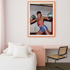 May include: A framed print showing a person in boxing gear. The person is wearing a red and blue outfit and green boxing gloves. The print is in an orange frame and is hung on a white wall.