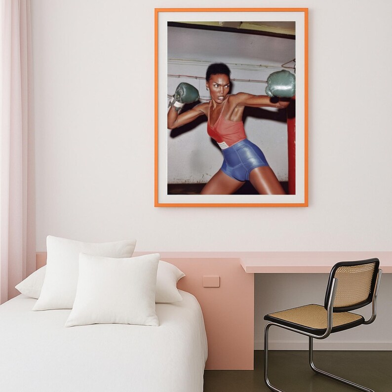 May include: A framed print showing a person in boxing gear. The person is wearing a red and blue outfit and green boxing gloves. The print is in an orange frame and is hung on a white wall.
