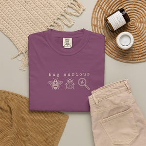 Op de afbeelding: Een pruimkleurig t-shirt met de witte tekst "bug curious" gecentreerd boven een lijntekening van een bij, een lieveheersbeestje en een vergrootglas. Het shirt heeft een ronde hals en een zichtbaar merklabel aan de binnenkant van de kraag.