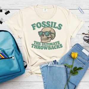 Peut inclure: T-shirt crème avec l'inscription "FOSSILS" au-dessus d'un crâne portant des lunettes de soleil. En dessous, le texte "THE ULTIMATE THROWBACKS" est en vert. Le t-shirt est associé à un jean bleu et un sac à dos turquoise.