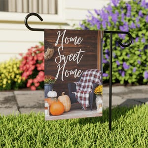 May include: A garden flag with the words "Home Sweet Home" in white script. The flag features a cozy porch scene with a chair, blanket, pumpkins, and flowers, evoking a warm autumn atmosphere.