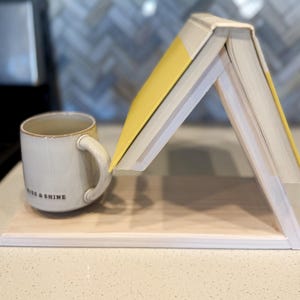 May include: A light-colored wooden book stand with a book and a mug. The book is open, with yellow pages visible. A white mug with the words "RISE & SHINE" is on the left. The stand is on a light-colored surface.